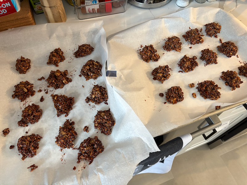 Two cookie sheets covered in parchment paper filled with twenty five blobs of a brown oat mixture. The mixture appears to be mostly comprised of oats, with a chocolatey semi-liquid filling the spaces between individual oats.